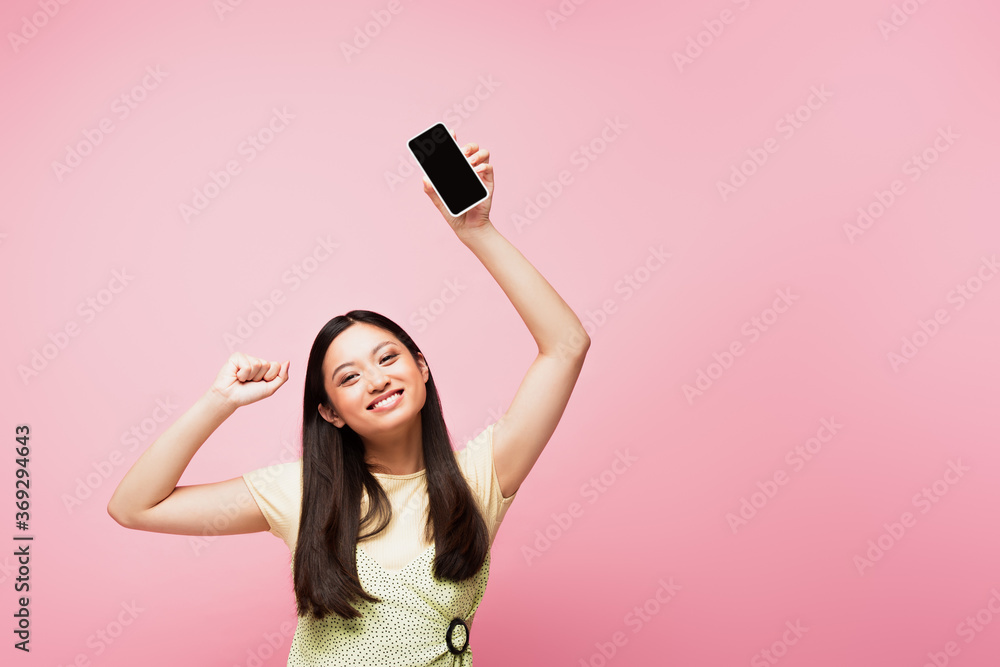 smiling asian girl with clenched fist holding smartphone with blank screen isolated on pink smiling asian girl with clenched fist holding smartphone with blank screen isolated on pink
