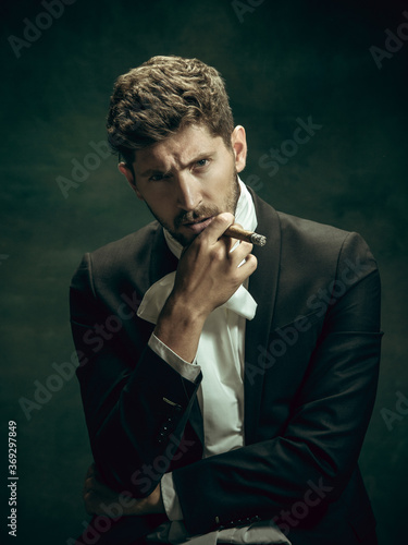 Brutal smokes cigar. Young man in suit as Dorian Gray isolated on dark green background. Retro style, comparison of eras concept. Beautiful male model like classic literature character, old-fashioned.