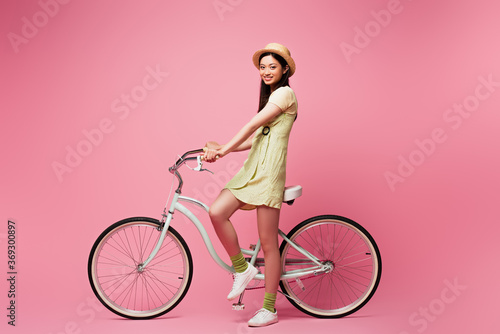 Fototapeta Naklejka Na Ścianę i Meble -  cheerful asian young woman in straw hat near bicycle on pink
