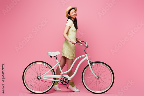 cheerful asian young woman in straw hat walking with bicycle on pink © LIGHTFIELD STUDIOS
