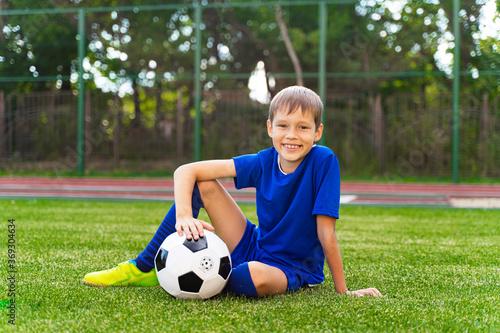 A little boy sits on a green soccer field, a soccer ball