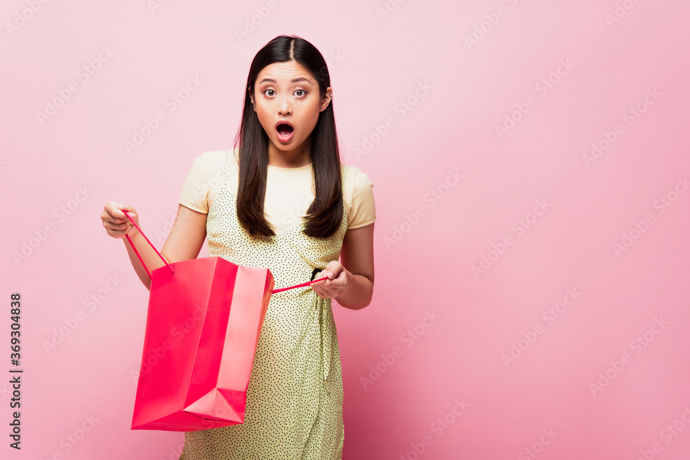 shocked young asian woman holding shopping bag on pink