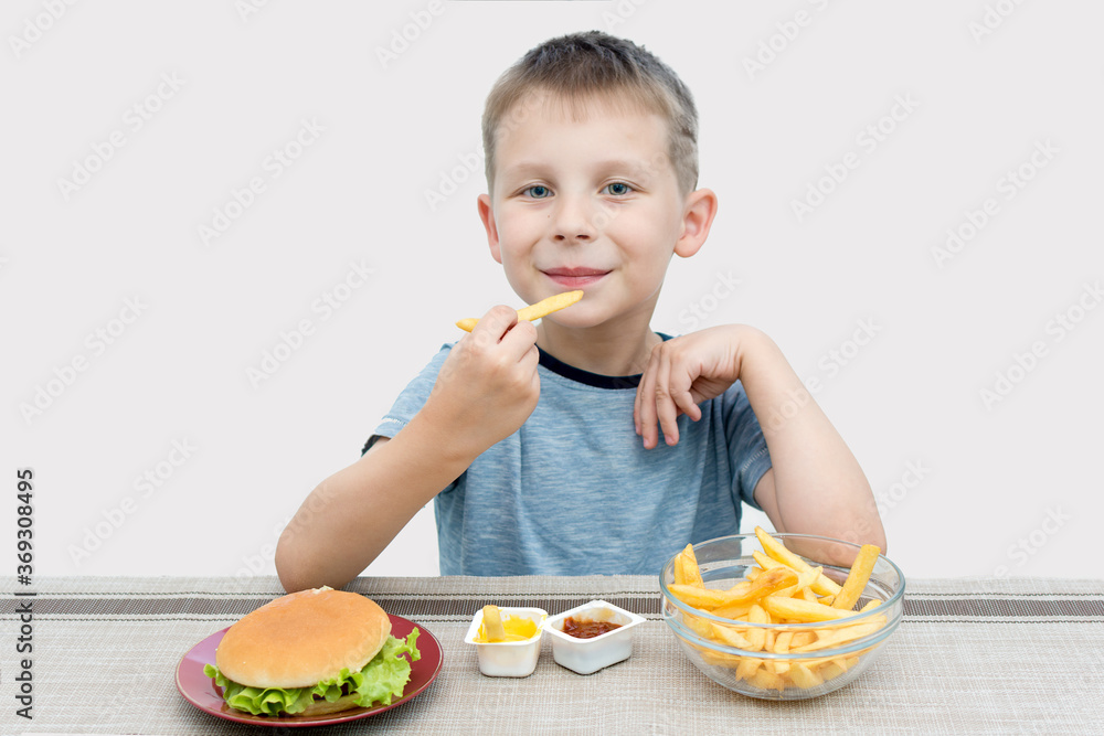 A small preschooler sits at the table and holds French fries in his hands. Fast food, hamburger, French fries, and gravy are on the table. The child eats food that is harmful to the growing body. A small preschooler sits at the table and holds French fries in his hands. Fast food, hamburger, French fries, and gravy are on the table. The child eats food that is harmful to the growing body.
