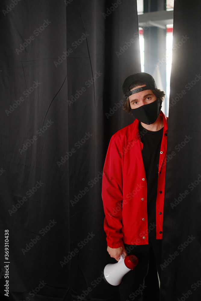 Emotional young man 25-30 years old in a black protective mask, a cap and a red jacket holding a megaphone against a dark background.