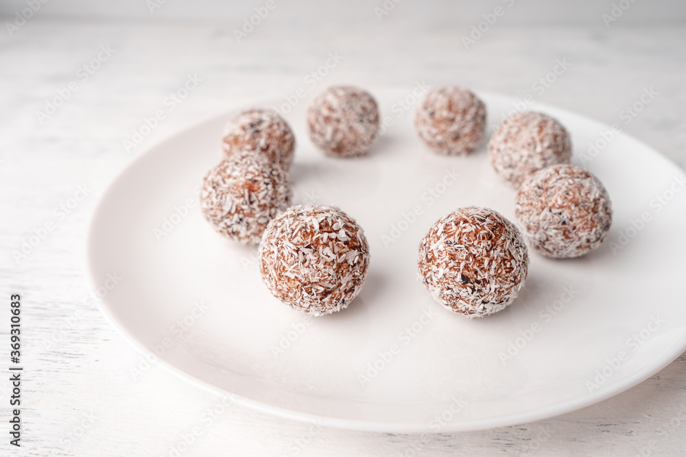 A white plate with round sweets made of dates and coconut shavings on a light wooden background A white plate with round sweets made of dates and coconut shavings on a light wooden background