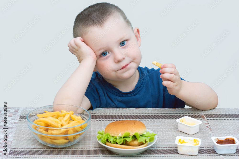 Little blue-eyed boy eating fast food. On a light background, the child is sad, does not smile. Concept - the child is tired of unhealthy, monotonous food, dreams of tasty, healthy, wholesome food.