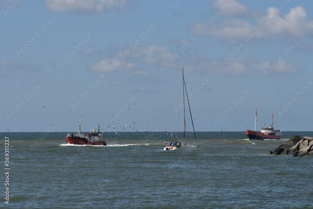 Zwei rote und eine weißer Schiff fahren auf das Meer. Unzählige Möwen fliegen umher . Auf der rechte Seite des Bildes sind noch große Betonsteine zu sehen. 