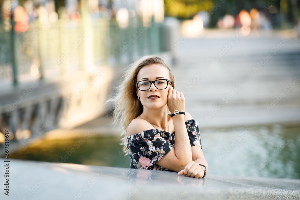 Beautiful young woman with blond curly hair wearing a dress and glasses in a park in the evening