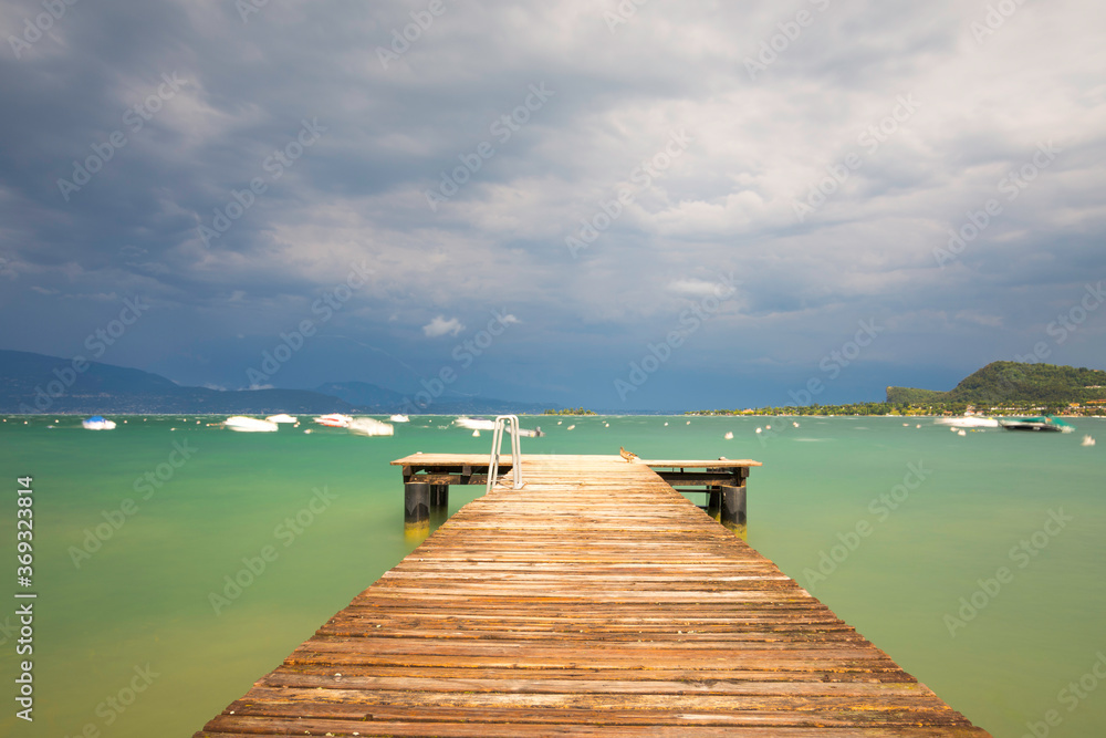 Fototapeta premium A footbridge leads to the troubled Lake Garda in Italy. The blue-green water surface is churned, lots of waves. Boats dance in the water. The sky is blue, a storm is coming. Long exposure time.