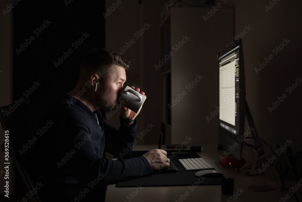 Man with earbuds drinking coffee and working late at computer in dark room Man with earbuds drinking coffee and working late at computer in dark room