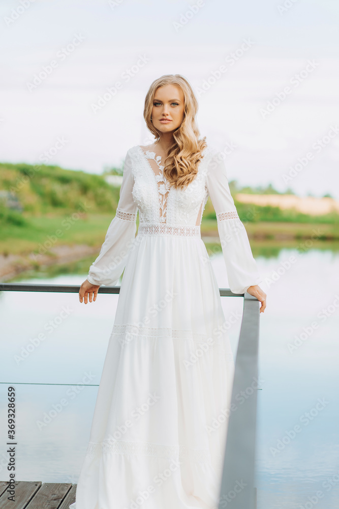 Blonde hair bride in white silk long lace dress standing on the terrace with lake view