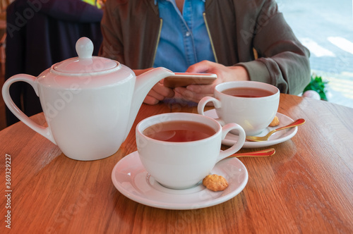 A teapot and two cups of tea on a table in a cafe. Tea for two. Young woman holding smartphone. Horizontal orientation, selective focus.