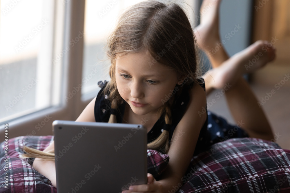 Smart small Caucasian girl lying on pillow look at tablet screen watch video online on gadget, little kid child have fun using modern pad device at home, play application game, technology concept Smart small Caucasian girl lying on pillow look at tablet screen watch video online on gadget, little kid child have fun using modern pad device at home, play application game, technology concept
