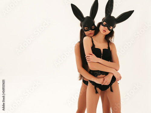 Two sexy beautiful women wearing carnival black mask of Easter bunny rabbit.Hot brunette girls posing near white wall in studio. Seductive models in nice lingerie