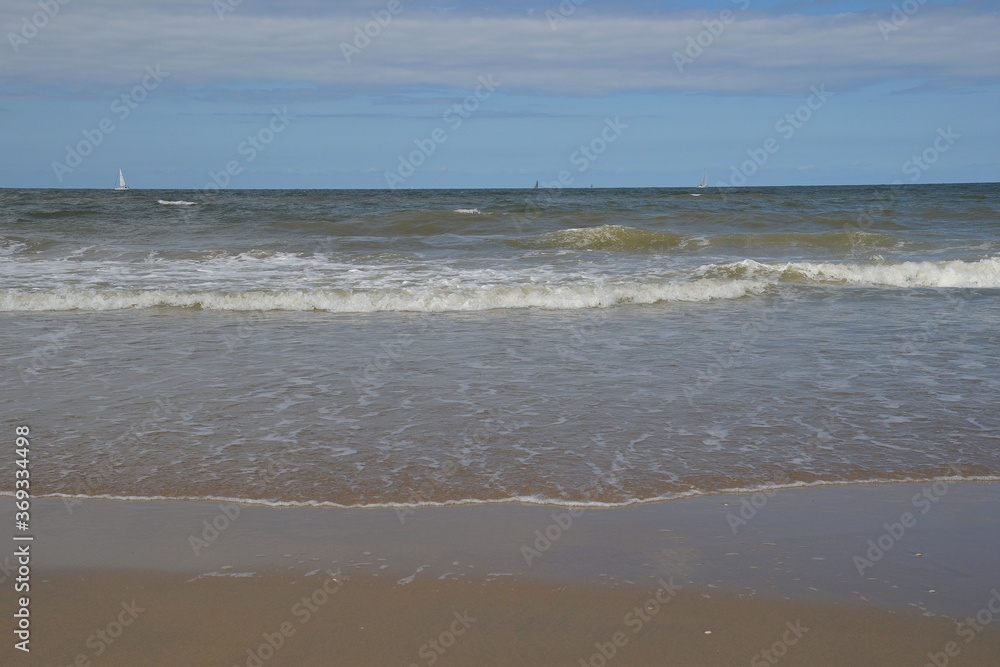 Das blaue Meer wird von mehreren weißen Wellen aufgewühlt. Am Horizont sieht man ganz klein mehrere Segelschiffe. 