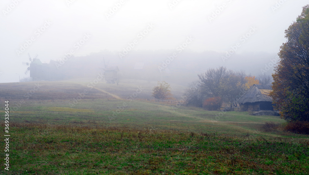 Foggy autumn landscape with a road on a hill in the middle of a field with windmills and a wooden hut.