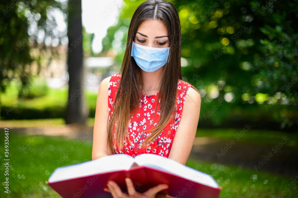 Beautiful female college student reading a book on a bench in a park and wearing a mask in coronavirus times - obrazy, fototapety, plakaty 