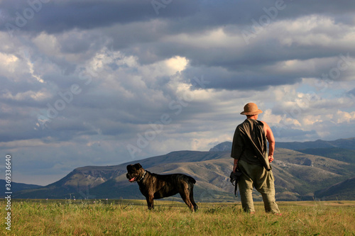 A woman in a hat with a dog of the Cane Corso breed stands on a meadow in a beautiful landscape with a view of the mountains and clouds on a summer day
