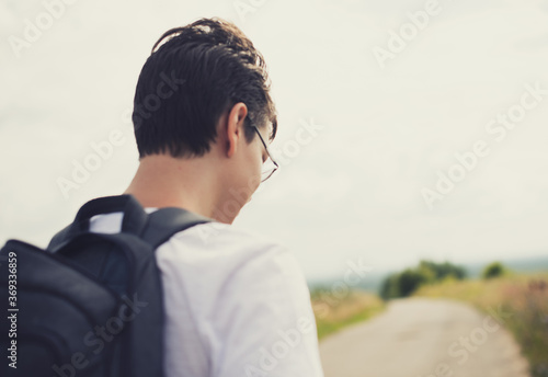 A teenager with glasses, a white t-shirt, and a backpack on a solo trip on the road to his goal in the fall.