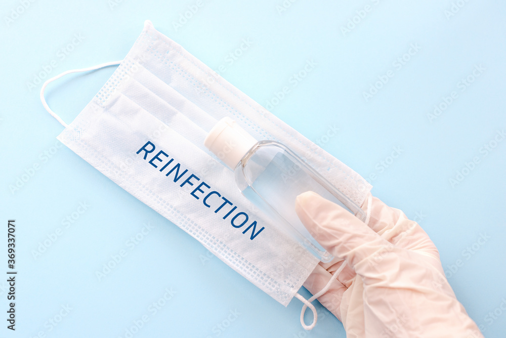 A hand in a protective glove holds a medical mask with the words REINFECTION and antiseptic, on a blue background. The concept of possible re-infection with the coronavirus covid-19. Flat lay A hand in a protective glove holds a medical mask with the words REINFECTION and antiseptic, on a blue background. The concept of possible re-infection with the coronavirus covid-19. Flat lay