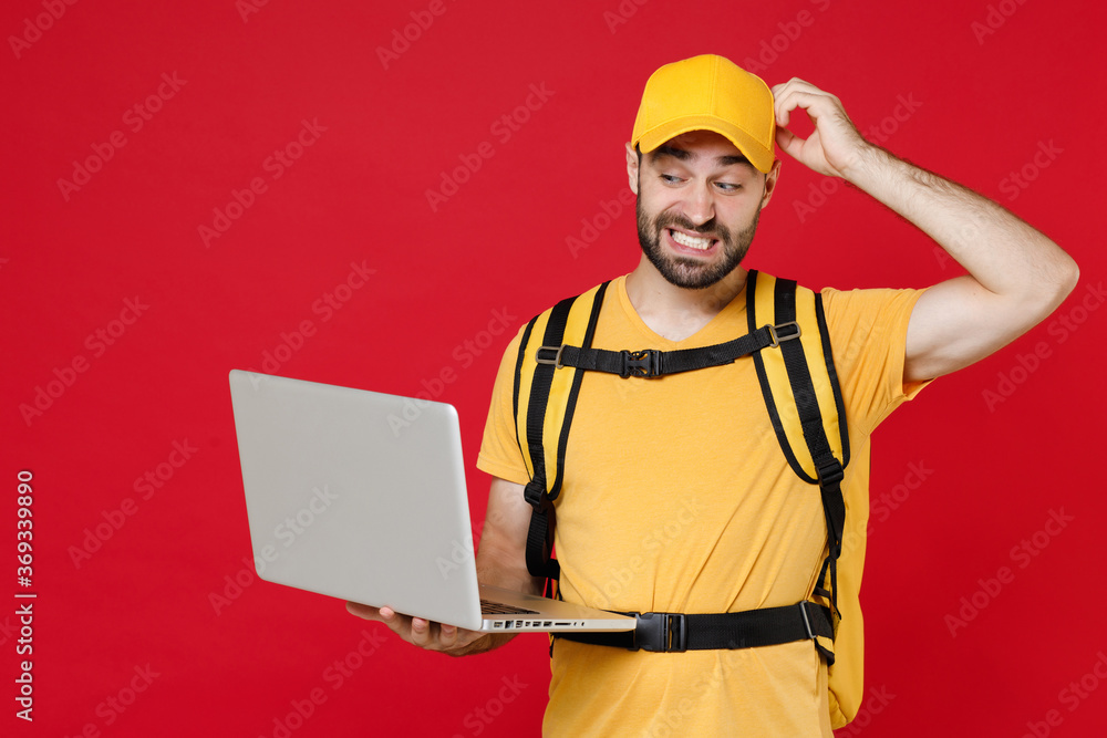 Delivery man in yellow cap t-shirt uniform thermal bag backpack with food hold laptop pc isolated on red background studio Guy male employee work as courier Service coronavirus covid-19 virus concept Delivery man in yellow cap t-shirt uniform thermal bag backpack with food hold laptop pc isolated on red background studio Guy male employee work as courier Service coronavirus covid-19 virus concept