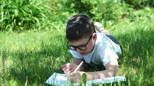portrait of studying schoolboy in glasses laying on grass writing in exercise book making homework. male child drawing geometric figure doing math outdoors. Elementary school education. Back to school