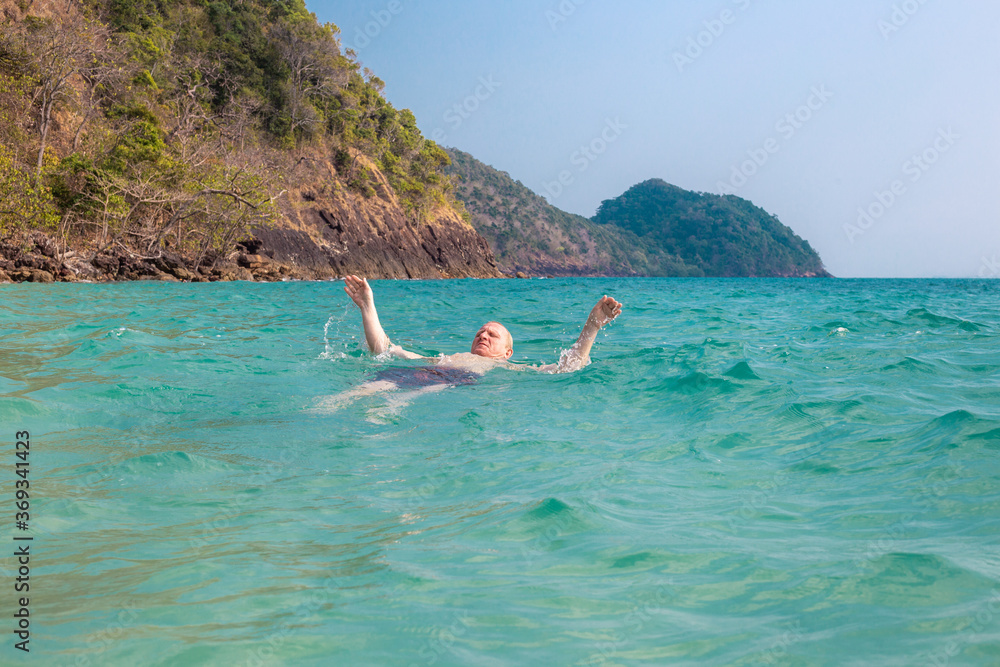 an adult man swims in the sea against the background of mountains, swims on his back, summer sea holidays, travel and tourism