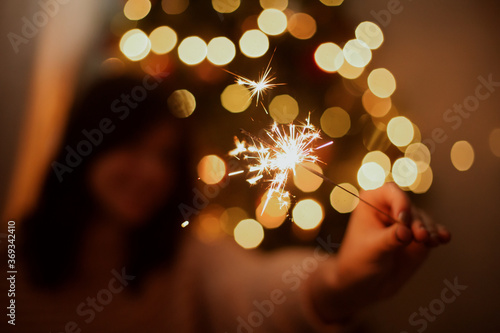 Stylish girl with burning sparkler celebrating in festive dark room. Happy New Year. Happy woman holding firework at christmas tree with golden lights.