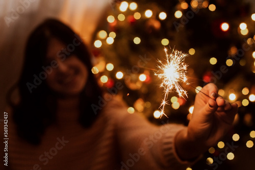 Happy woman holding firework at christmas tree with golden lights. Stylish girl with burning sparkler celebrating in festive dark room. Happy New Year