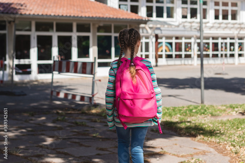 A girl with a pink satchel on her back approaches the school door. The first day of school, the children are ready to study after the holidays. Education, lessons after the pandemic