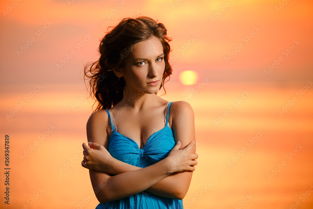 Young romantic girl walks relaxed by the water at sunset or sunrise. Beautiful woman in blue dress in boho style on background at sea. Sunrise over the ocean. Summertime scene Young romantic girl walks relaxed by the water at sunset or sunrise. Beautiful woman in blue dress in boho style on background at sea. Sunrise over the ocean. Summertime scene