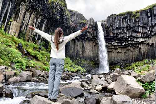 Iceland. Woman enjoying majestic Svartifoss waterfall. Female is visiting famous tourist attraction of Iceland. Spectacular natural landmark on vacation in Skaftafell. Icelandic nature landscape.