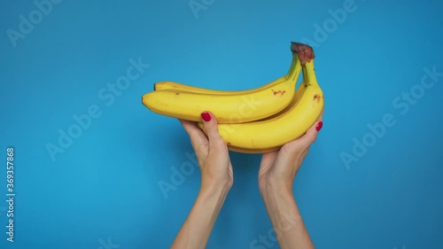 Woman hands with raises bunch bananas on blue background. Close up hands shows bananas, twists fruits