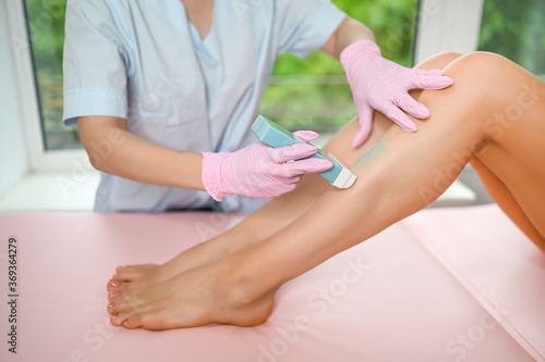 Woman with long tanned perfect legs and smooth skin having wax stripe depilation hair removal procedure on legs in beauty salon. Beautician in blue robe  pink gloves. Body care  epilation spa concept