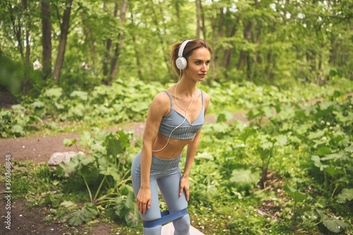 Young happy beautiful slim fit girl in blue sportswear with big white headphones doing squats exercise with fitness training gum in the summer green park outside. Girl with perfect body, sport yoga.