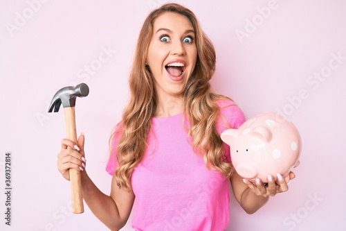 Young caucasian woman with blond hair holding piggy bank and hammer celebrating crazy and amazed for success with open eyes screaming excited.