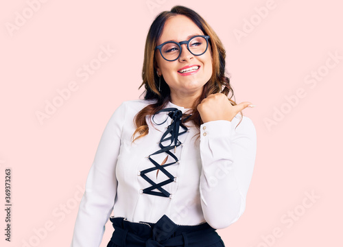 Young beautiful caucasian woman wearing business shirt and glasses smiling with happy face looking and pointing to the side with thumb up. photo