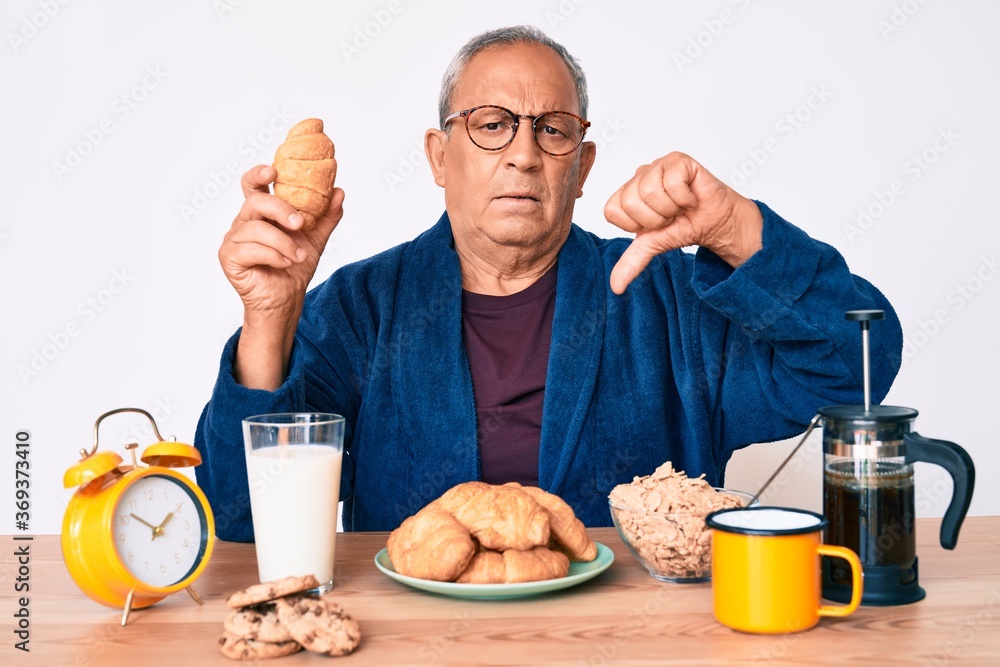 Senior handsome man with gray hair sitting on the table eating croissant for breakfast with angry face, negative sign showing dislike with thumbs down, rejection concept Senior handsome man with gray hair sitting on the table eating croissant for breakfast with angry face, negative sign showing dislike with thumbs down, rejection concept