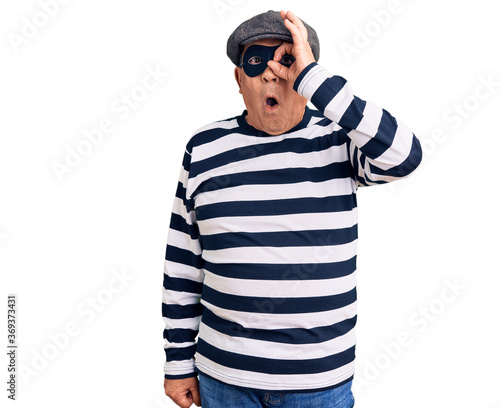 Senior handsome man wearing burglar mask and t-shirt doing ok gesture shocked with surprised face, eye looking through fingers. unbelieving expression.