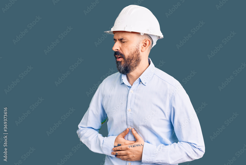 Young handsome man wearing architect hardhat with hand on stomach because nausea, painful disease feeling unwell. ache concept. Young handsome man wearing architect hardhat with hand on stomach because nausea, painful disease feeling unwell. ache concept.