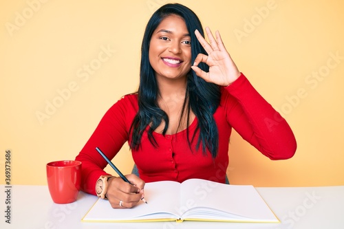 Beautiful latin young woman with long hair writing a book sitting on the table doing ok sign with fingers, smiling friendly gesturing excellent symbol