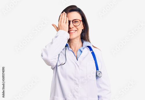 Young beautiful woman wearing doctor stethoscope and glasses covering one eye with hand, confident smile on face and surprise emotion.