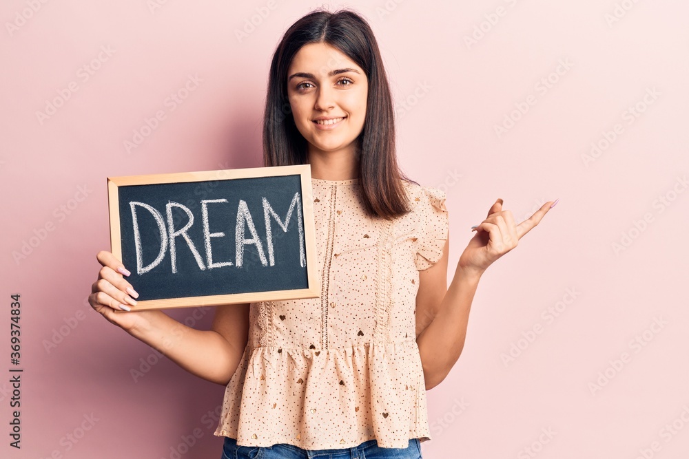Young beautiful girl holding blackboard with dream word message smiling happy pointing with hand and finger to the side Young beautiful girl holding blackboard with dream word message smiling happy pointing with hand and finger to the side