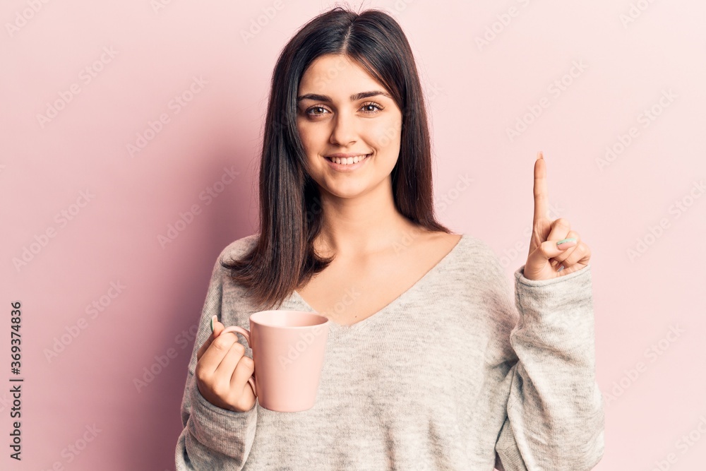 Young beautiful girl drinking mug of coffee smiling with an idea or question pointing finger with happy face, number one Young beautiful girl drinking mug of coffee smiling with an idea or question pointing finger with happy face, number one