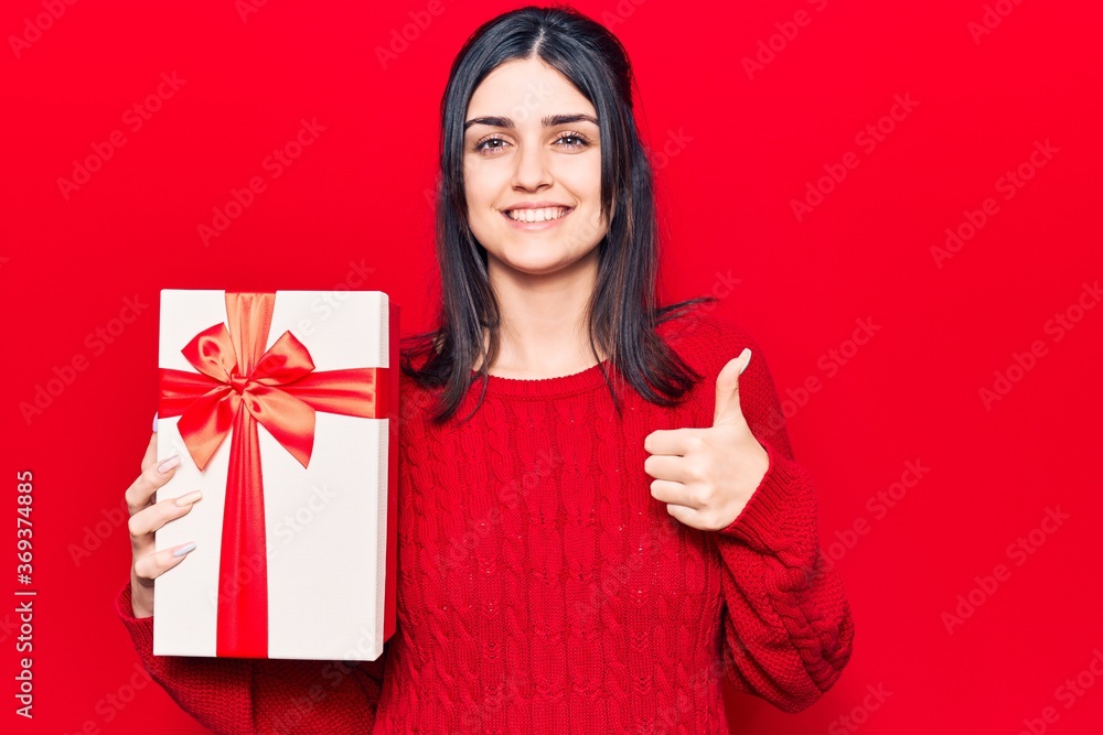Young beautiful girl holding gift smiling happy and positive, thumb up doing excellent and approval sign Young beautiful girl holding gift smiling happy and positive, thumb up doing excellent and approval sign