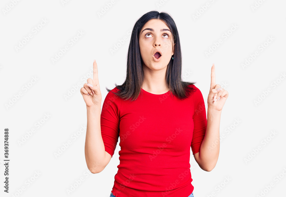 Young beautiful girl wearing casual t shirt amazed and surprised looking up and pointing with fingers and raised arms. Young beautiful girl wearing casual t shirt amazed and surprised looking up and pointing with fingers and raised arms.