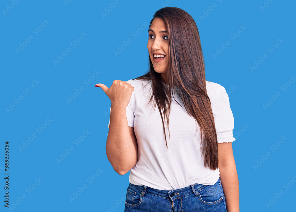 Young beautiful brunette woman wearing casual t-shirt smiling with happy face looking and pointing to the side with thumb up. Young beautiful brunette woman wearing casual t-shirt smiling with happy face looking and pointing to the side with thumb up.