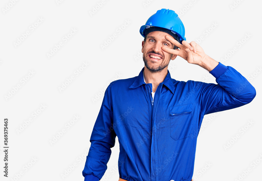 Young handsome man wearing worker uniform and hardhat doing peace symbol with fingers over face, smiling cheerful showing victory Young handsome man wearing worker uniform and hardhat doing peace symbol with fingers over face, smiling cheerful showing victory