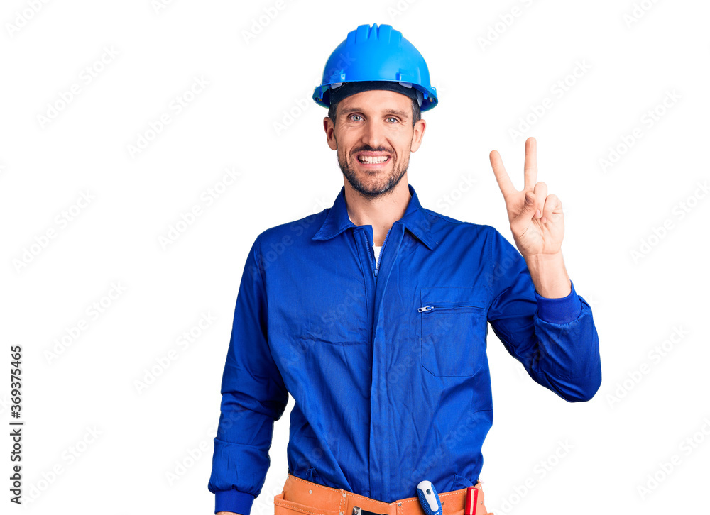 Young handsome man wearing worker uniform and hardhat showing and pointing up with fingers number two while smiling confident and happy.