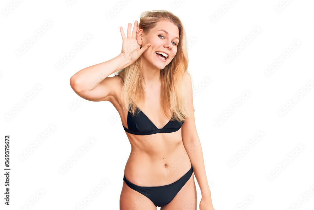 Young beautiful blonde woman wearing bikini smiling with hand over ear listening an hearing to rumor or gossip. deafness concept. Young beautiful blonde woman wearing bikini smiling with hand over ear listening an hearing to rumor or gossip. deafness concept.